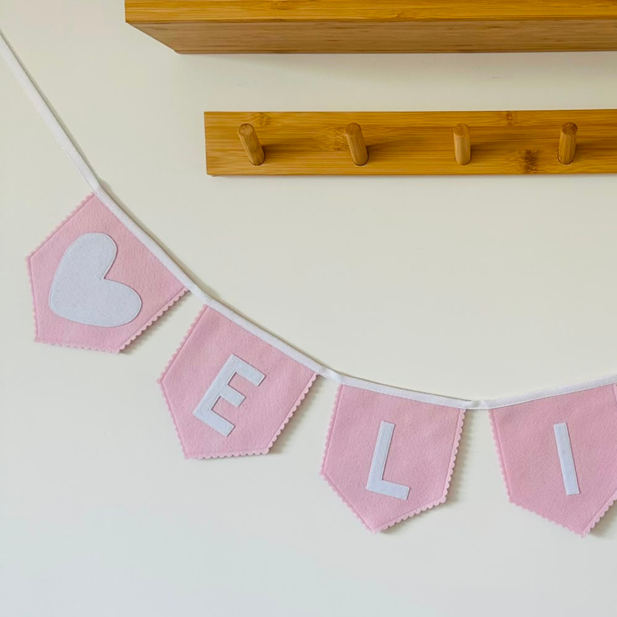 Close-up van een handgemaakte licht roze vilten hart vlaggenlijn met naam, met een wit vilten hart-icoon en de letters "F", "L" en "I" in wit vilt.