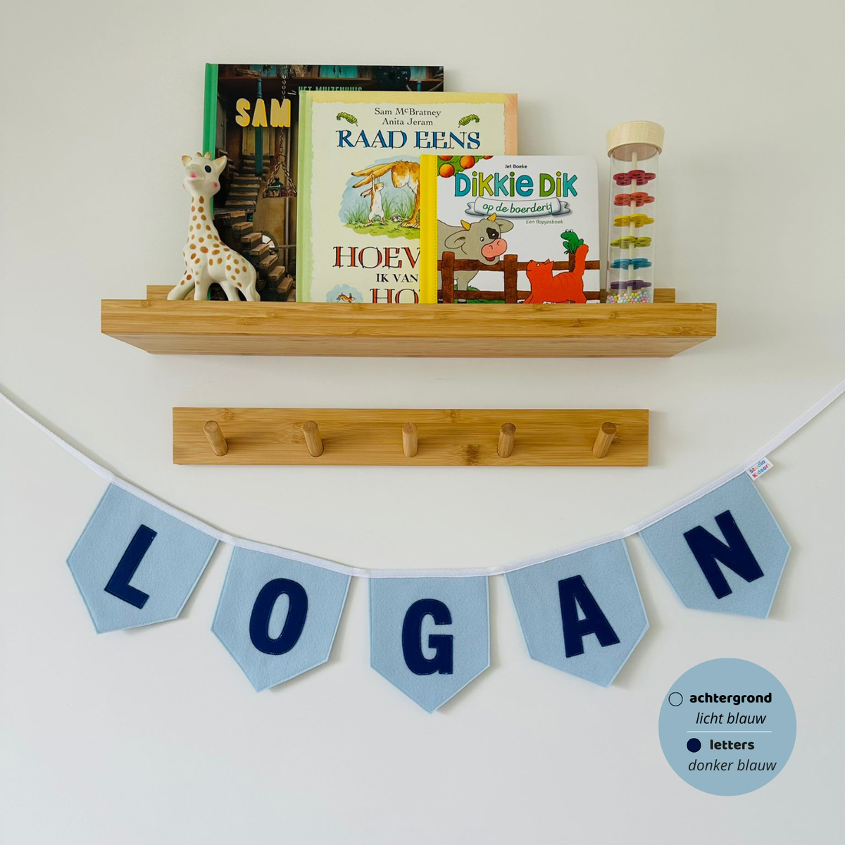 Gepersonaliseerde handgemaakte vilten vlaggenlijn met de naam "LOGAN", bestaande uit licht blauw kleurige vlaggen met donker blauw vilten letters. De randen van de vlaggen zijn afgewerkt met een rechte rand. De vlaggenlijn hangt aan een naturel katoenen lint in een kinderkamer, gefotografeerd onder een houten boekenplank met kinderboeken en een Sophie de Giraf speeltje, boven een houten kapstok.