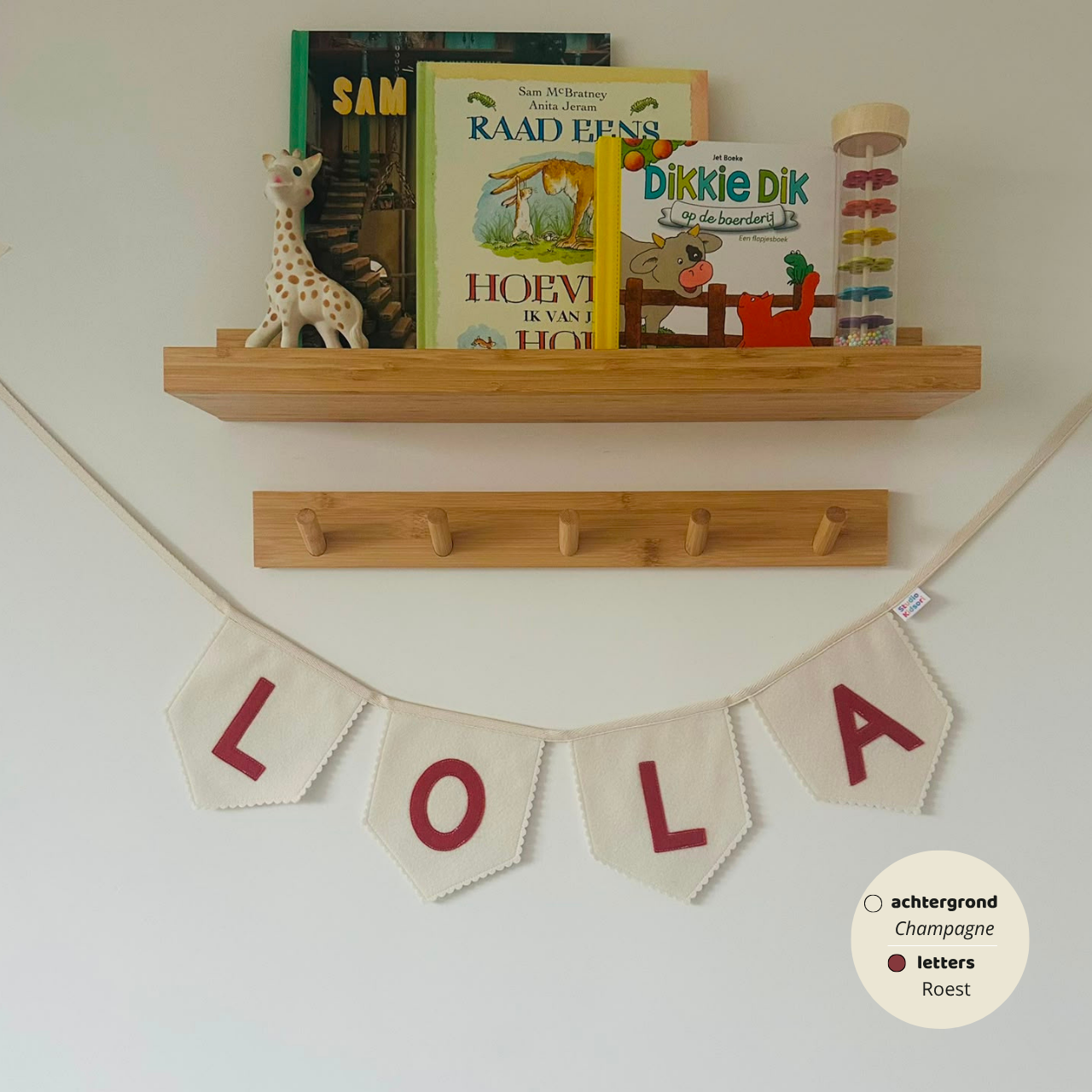 Gepersonaliseerde handgemaakte vilten vlaggenlijn met de naam "LOLA", bestaande uit champagne kleurige vlaggen met roest vilten letters. De randen van de vlaggen zijn afgewerkt met een scallop rand. De vlaggenlijn hangt aan een naturel katoenen lint in een kinderkamer, gefotografeerd onder een houten boekenplank met kinderboeken en een Sophie de Giraf speeltje, boven een houten kapstok.
