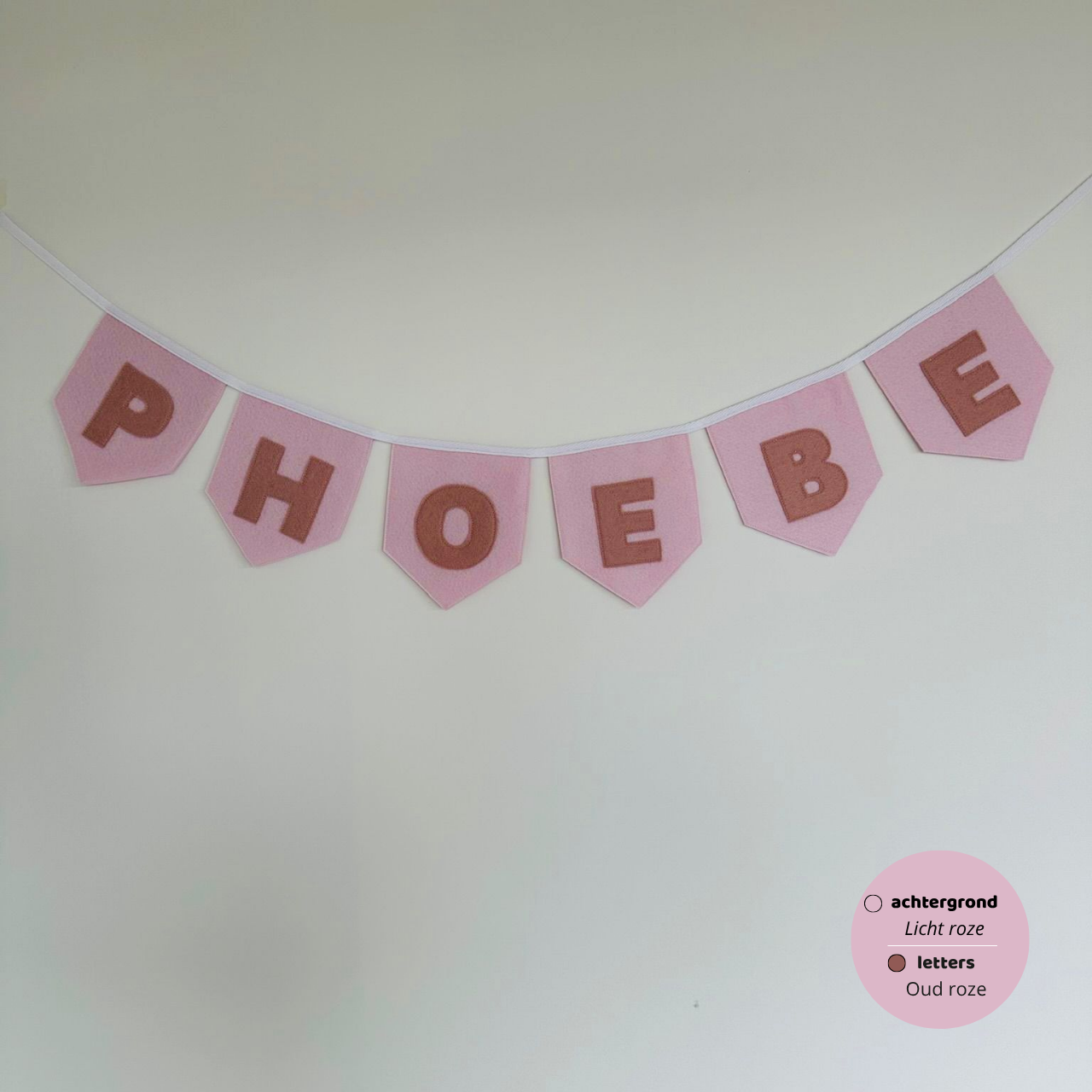 Gepersonaliseerde handgemaakte vilten vlaggenlijn met de naam "Phoebe", bestaande uit licht licht roze vlaggen met oud  oze vilten letters. De randen van de vlaggen zijn afgewerkt met een rechte rand. De vlaggenlijn hangt aan een naturel katoenen lint.