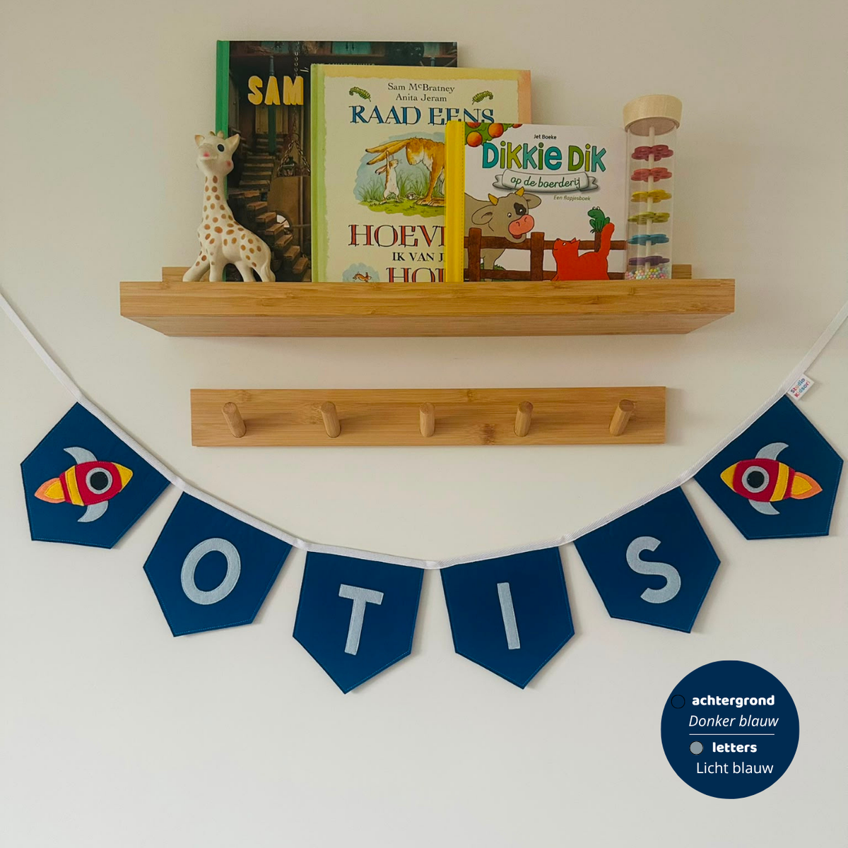 Gepersonaliseerde handgemaakte vilten naamvlaggenlijn met de naam "OTIS", bestaande uit lichtblauw kleurige vilten letters met donkerblauwe vlaggen met een raket op het eerste vlaggetje en het laatste vlaggetje. De randen van de vlaggen zijn afgewerkt met een rechte afwerking. De vlaggenlijn hangt aan een wit katoenen lint.
