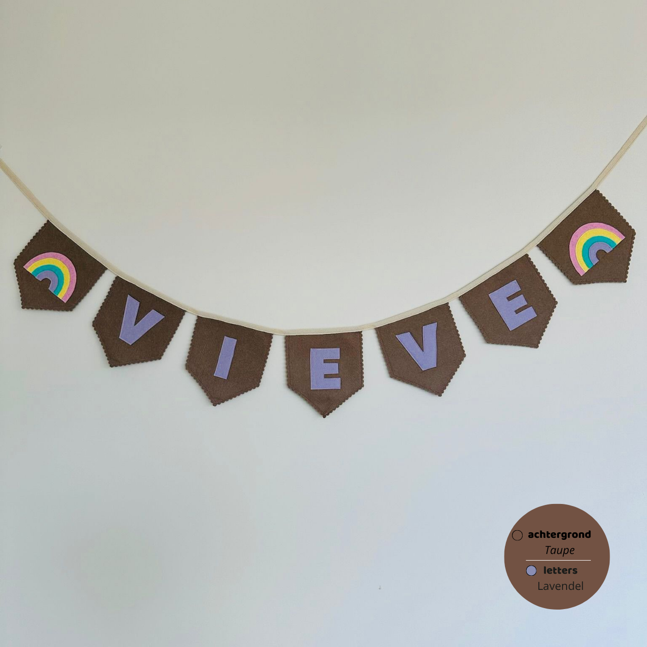 Gepersonaliseerde handgemaakte vilten naamvlaggenlijn met de naam "Vieve", bestaande uit Taupe kleurige vlaggen met lavendel kleurige vilten letters met een regenboog het eerste vlaggetje en het laatste vlaggetje. De randen van de vlaggen zijn afgewerkt met een scalp afwerking. De vlaggenlijn hangt aan een katoenen lint.