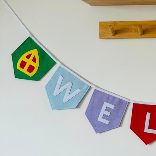 Sinterklaas vlaggenlijn van vilt met de letters WEL in witte letters op gekleurde vlaggetjes, met aan de uiteinden een mijter en een Pietenmuts, hangend aan een muur boven een houten wandplank
