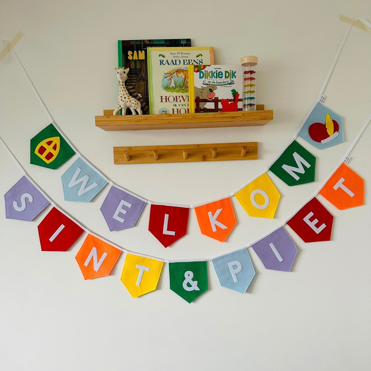 Sinterklaas vlaggenlijn van vilt met de letters ''welkom sint & piet''  in witte letters op gekleurde vlaggetjes, met aan de uiteinden een mijter en een Pietenmuts bij welkom, hangend aan een muur boven een houten wandplank