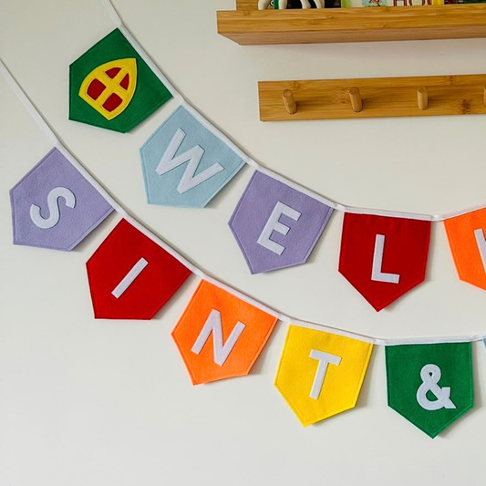 Sinterklaas vlaggenlijn van vilt met de letters ''wel sint &''  in witte letters op gekleurde vlaggetjes, met aan de uiteinden een mijter en een Pietenmuts bij welkom, hangend aan een muur boven een houten wandplank