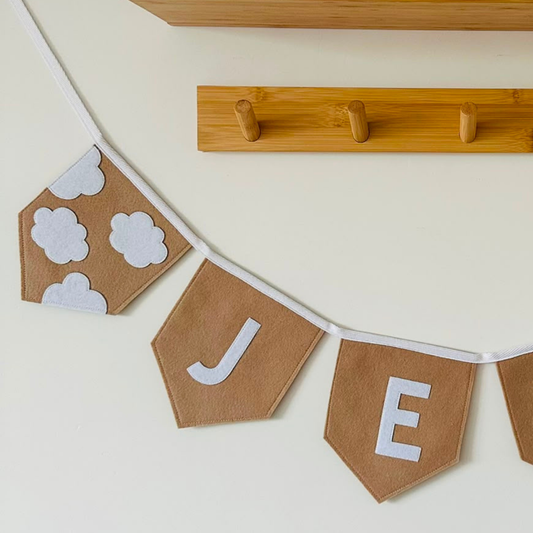 Close-up van een handgemaakte vilten wolken vlaggenlijn met naam, beige vlaggetjes met witte vilten wolkjes en de letters "J" en "E", opgehangen aan een wit katoenen koord voor een houten wandkapstok.